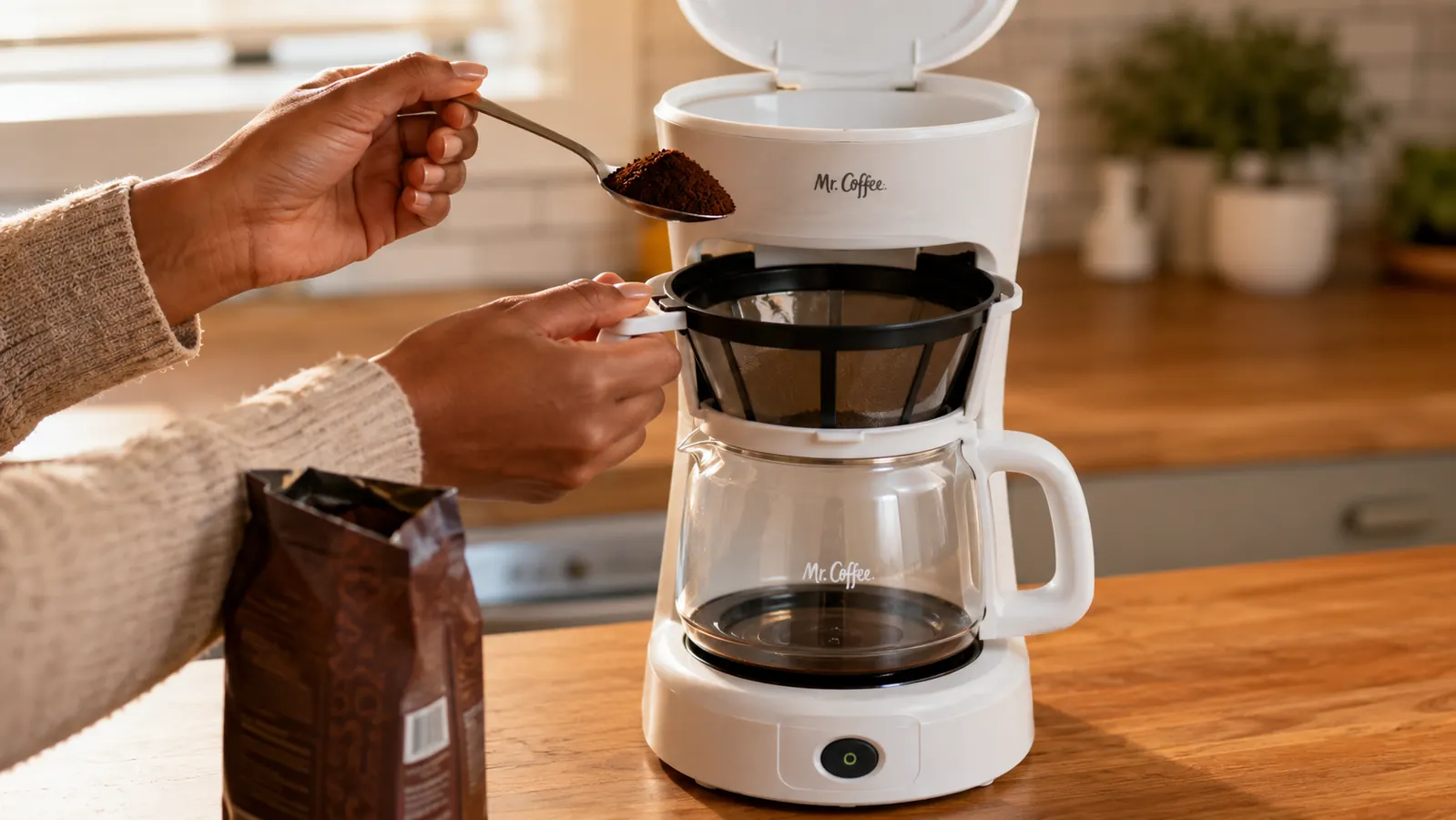 Hand spooning ground coffee from a bag into a drip coffee maker basket, reusable filter visible