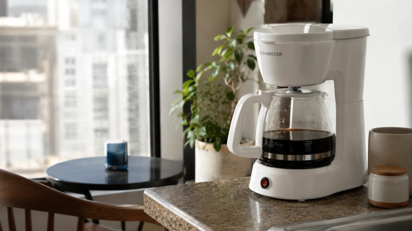 Compact white drip coffee maker on a small apartment kitchen counter next to a full glass carafe of fresh coffee
