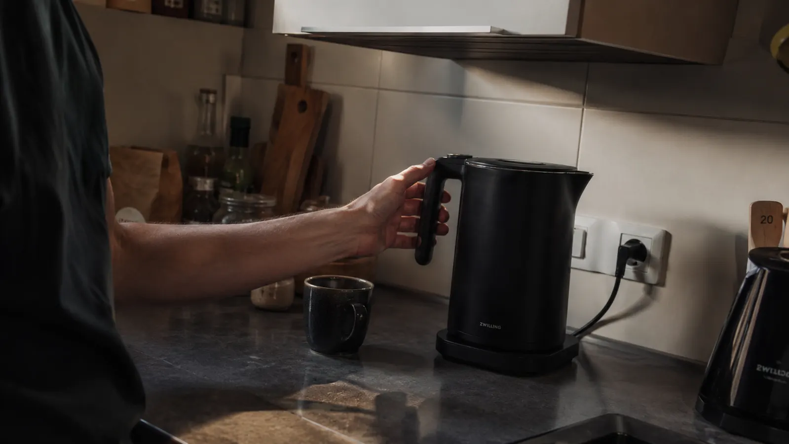 Cosori black electric kettle on a small kitchen counter with a hand reaching for the handle, overhead cabinet visible above