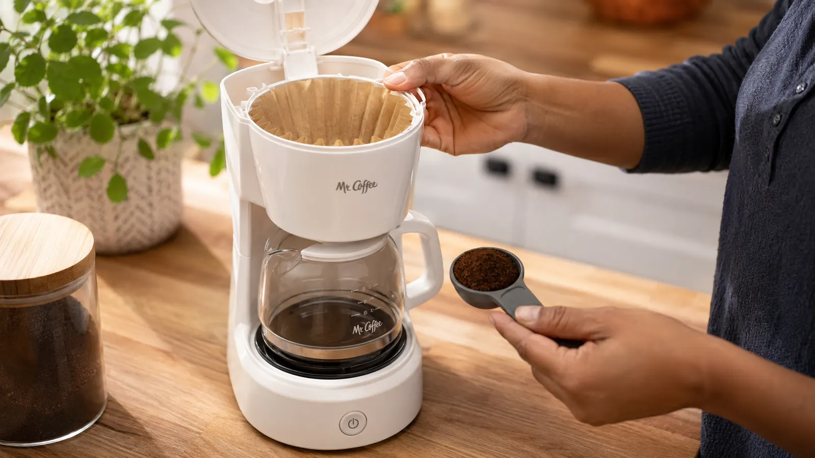A paper cone filter folded and seated inside the filter basket of a small coffee maker, with ground coffee at the right fill level shown with a measuring spoon