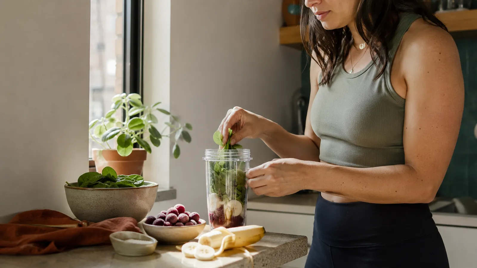 A compact personal blender cup on a small apartment kitchen counter next to fresh spinach, a banana, and a handful of frozen berries